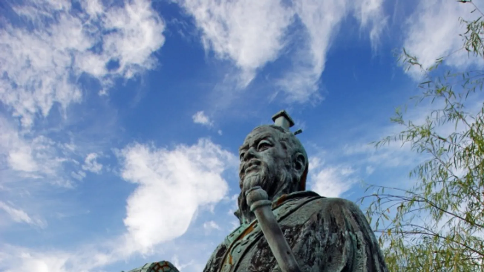 A monumental bronze statue of Sun Tzu holding a bamboo scroll, photographed in profile at the Enchoen garden in Yurihama, Japan.